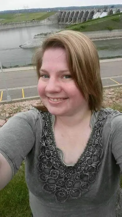Alicia Hummel taking a selfie on a grassy slope, in a gray top with lace trim, with a river and a dam behind her