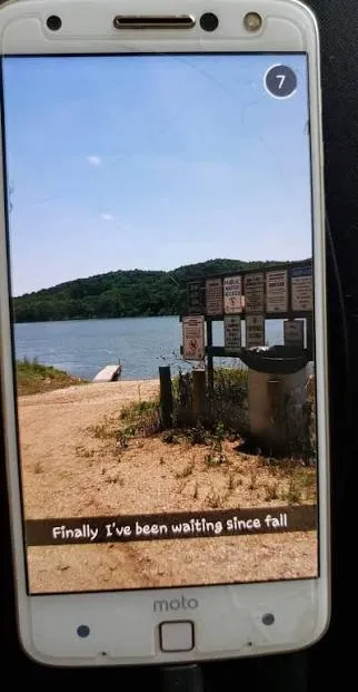 A Snapchat on a phone screen showing a dirt boat ramp, a sign board and a dock on the river, with the caption ‘Finally I’ve been waiting since fall’