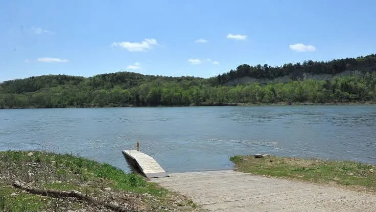 A concrete boat ramp leading to a small dock on a wide river, with wooded bluffs on the far bank
