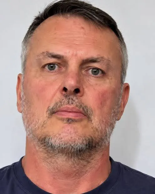 Kelly Grant, with short graying hair and a gray beard, in a navy T-shirt against a white wall