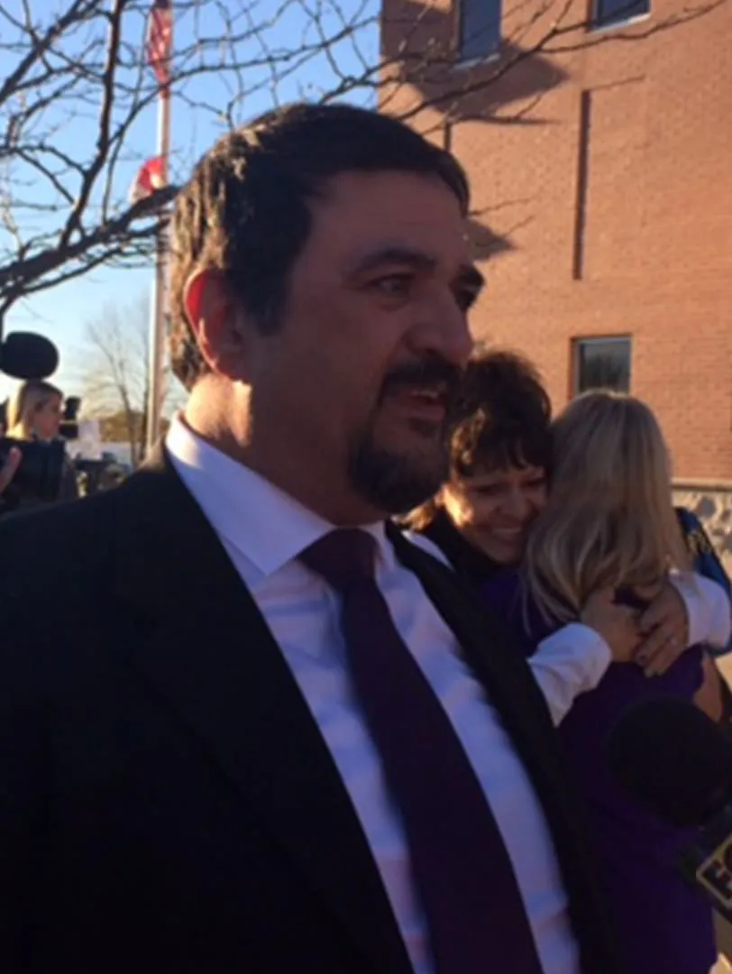 Russ Faria in a suit and tie outside a brick courthouse, with reporters’ microphones nearby