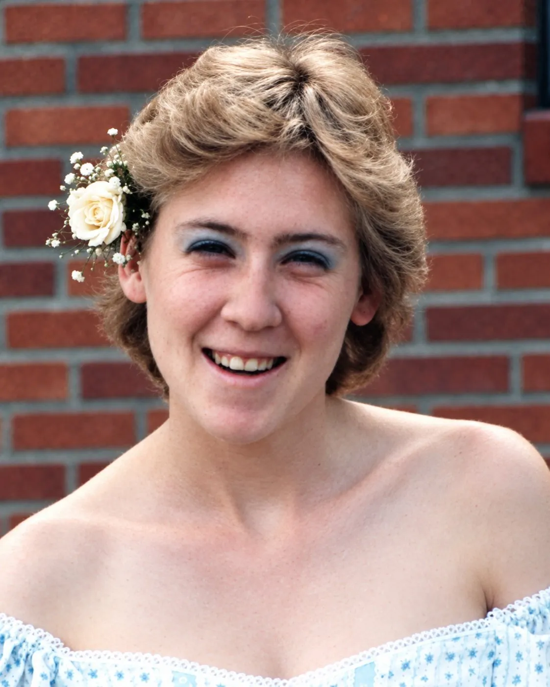 Carla Anderson laughing, with short hair and a white rose tucked behind her ear, in front of a red brick wall