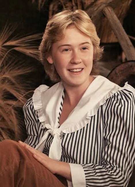 Carla Anderson smiling, with short blonde hair, in a striped blouse with a wide white ruffled collar