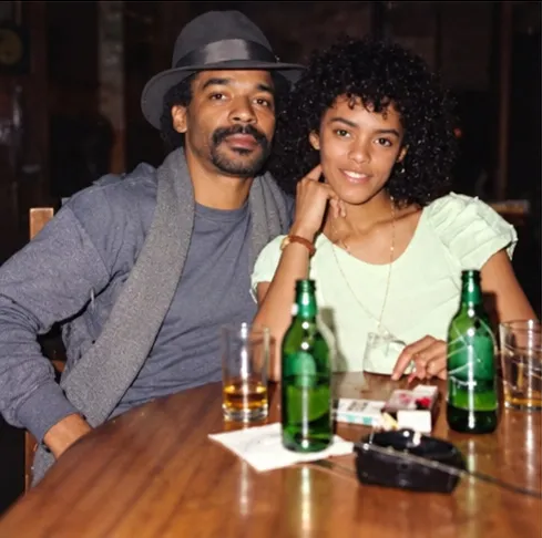 Cassandra Rhines smiling, her chin on her hand, seated at a wooden table beside a man in a gray hat and scarf, with two green bottles in front of them