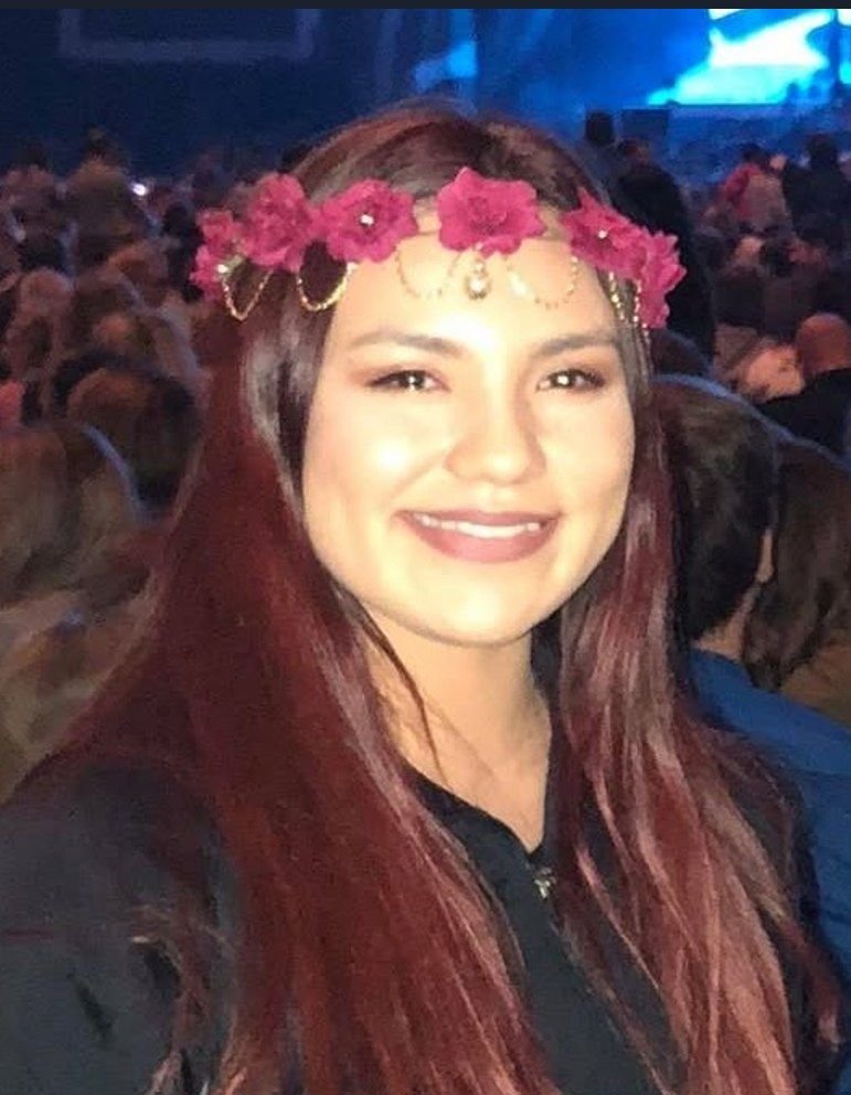 Kara Greger smiling, with long dark red hair and a crown of pink flowers, in a crowd at an indoor event