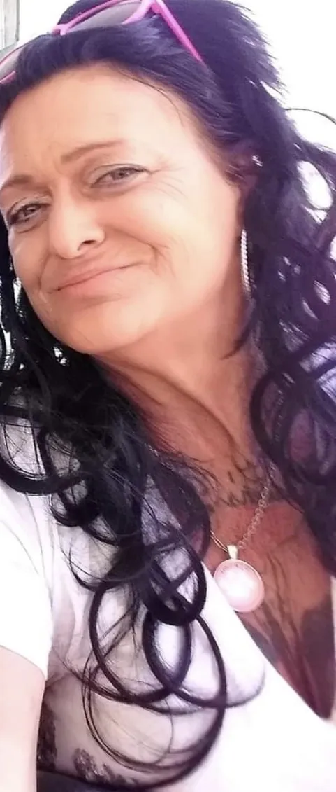 Linda Dillard, smiling, with long dark curly hair, pink sunglasses pushed up on her head and a pendant necklace