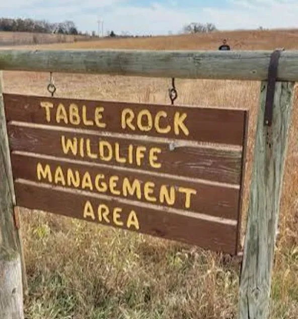 A brown wooden sign with yellow letters reading Table Rock Wildlife Management Area, hanging from a wooden post in front of a field of dry grass