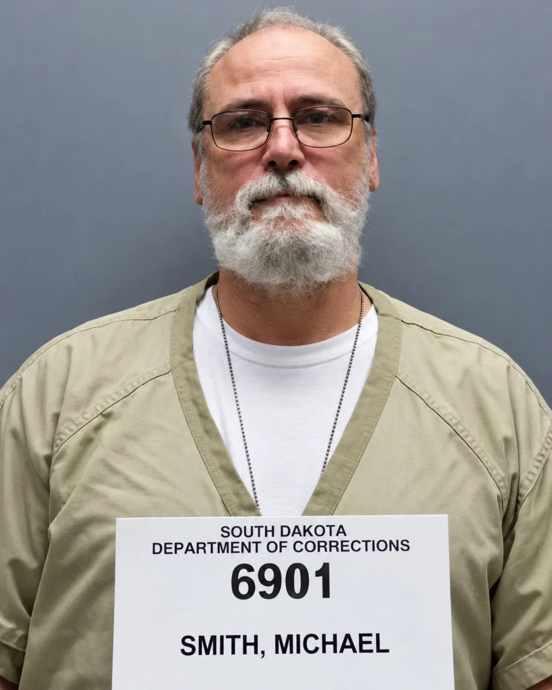 Michael Lee Smith, older, with a white beard and glasses, above a South Dakota Department of Corrections booking board