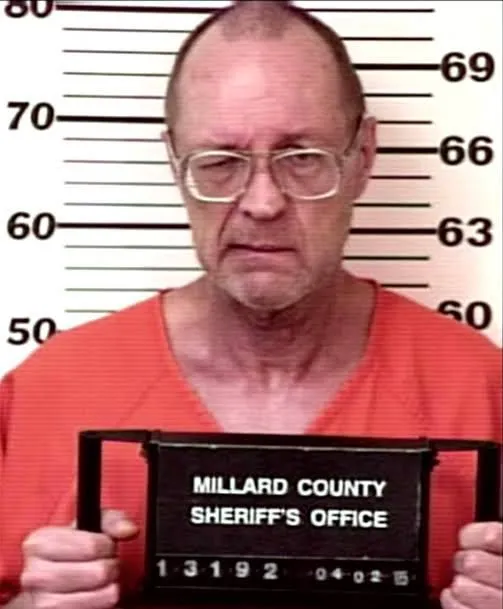 Booking photo of Robert Ben Rhoades, bald and wearing glasses and an orange jumpsuit, holding a Millard County Sheriff’s Office placard dated April 2, 2005