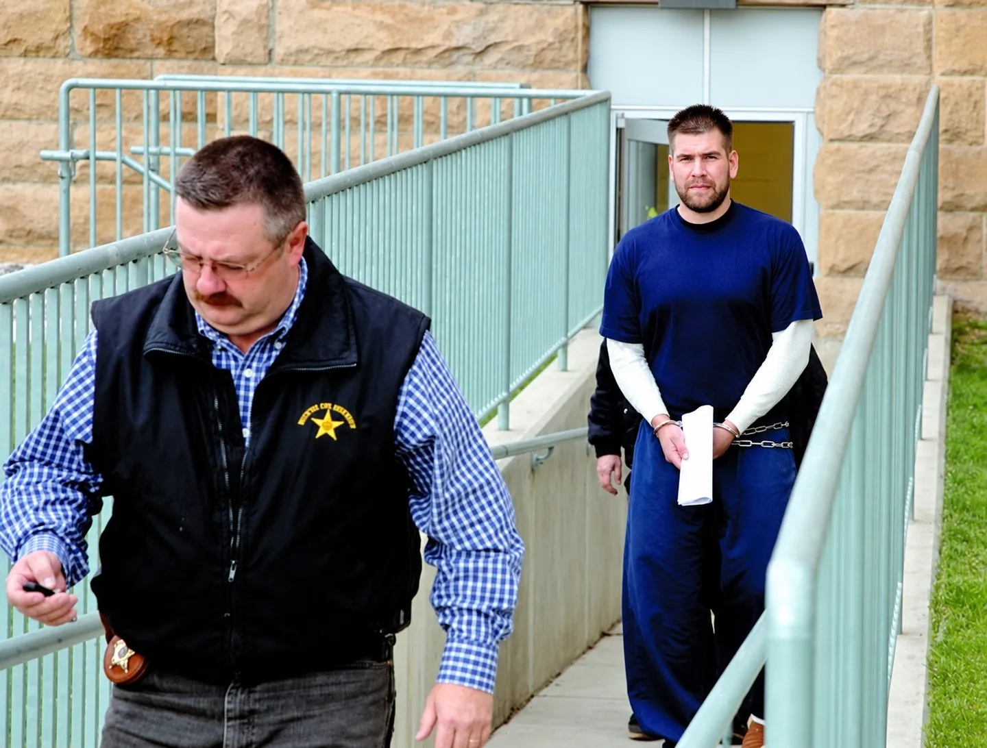 A man in a navy T-shirt over a white long-sleeved shirt, in handcuffs and a belly chain, walking down a ramp behind a sheriff’s officer in a black vest