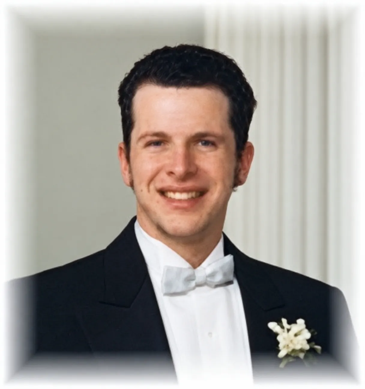 Troy Klug smiling, with short dark hair, in a black tuxedo, white bow tie and a white boutonniere
