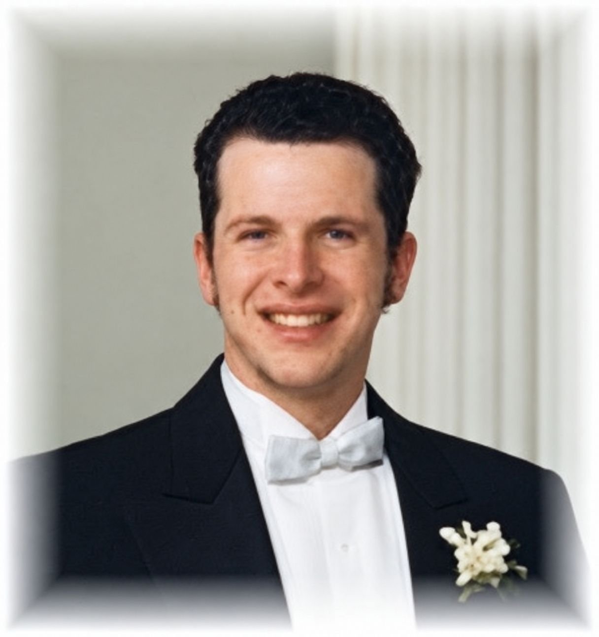 Troy Klug smiling, with short dark hair, in a black tuxedo, white bow tie and a white boutonniere