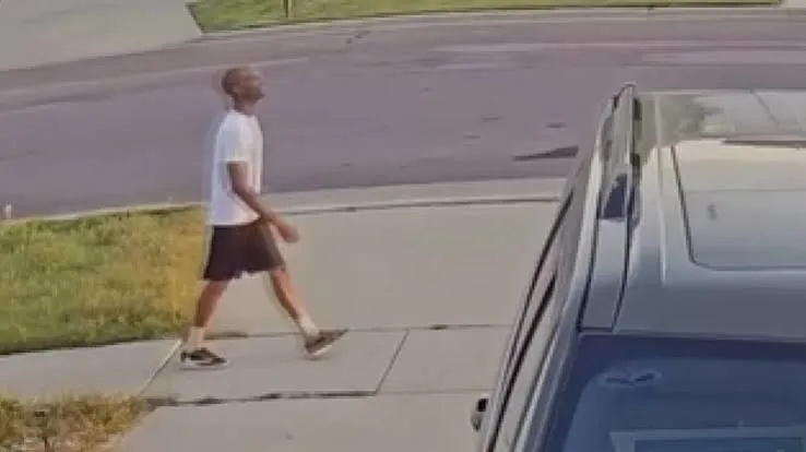 A man in a white T-shirt, black shorts and dark sneakers walking along a residential sidewalk, seen from above near a parked car